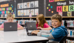 Three students with laptops sit at a table together. The student in the foreground is smiling at the camera