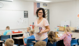 A teacher stands in a classroom holding books while elementary school students read at their desks